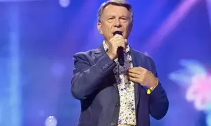 Wojciech Gąsowski w niebieskiej marynarce i kwiecistej koszuli śpiewa do mikrofonu na tle niebieskiej sceny.