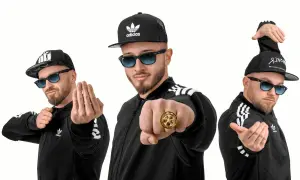 Trzech mężczyzn w czarnych bluzach z paskami Adidas i czapkach z daszkiem, pozujących jak zespoły disco polo.
