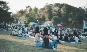 Dwie dziewczyny siedzą na trawie, ciesząc się atmosferą festiwalu. W tle widać tłum ludzi i kolorowe dekoracje, zapowiadające wspaniałe igo koncerty.