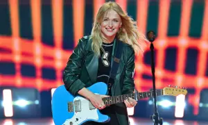 Uśmiechnięta polska piosenkarka z blond włosami gra na niebieskiej gitarze elektrycznej na tle pomarańczowych świateł.