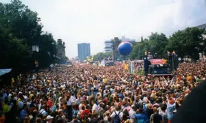 Tłum ludzi na ulicy podczas Love Parade, z wielkim niebieskim balonem i ciężarówką z platformą.