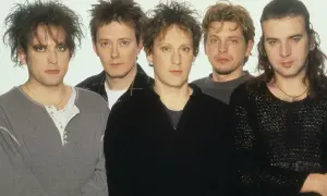Pięciu mężczyzn, członkowie zespołu The Cure, pozuje do zdjęcia. Ich charakterystyczne fryzury i makijaż podkreślają mroczny klimat ich muzyki.