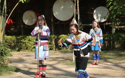 Trzy dziewczyny w barwnych strojach ludowych, jedna trzyma kij, druga instrument, w tle wielkie bębny.