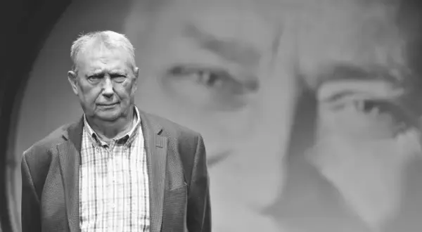 Wojciech Młynarski, jakby z tekstu "Jesteśmy na wczasach", w okryciu w kratę, na tle swojego portretu.