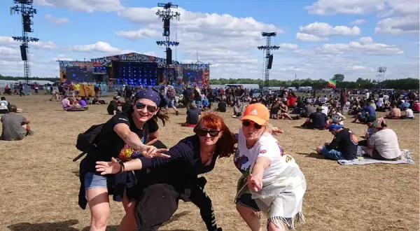 Trzy kobiety pozują na tle sceny festiwalowej Pol'and'Rock. Tłum ludzi siedzi na trawie, ciesząc się pogodą i oczekując na występ.