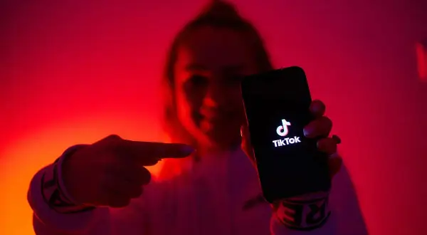 Dłoń wskazuje na ekran telefonu z logo TikToka. W tle czerwone, neonowe światła, tworzące atmosferę jak z klubu z tantiemami.