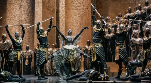 Scena z opery Aida: tancerze w czarnych strojach ze złotymi zdobieniami i maskami na twarzach, niektórzy z włóczniami, inni leżą na scenie.