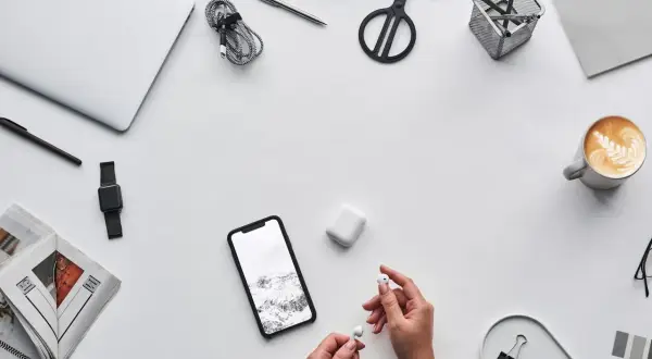 Dłonie trzymające białe słuchawki AirPods obok smartfona i etui.