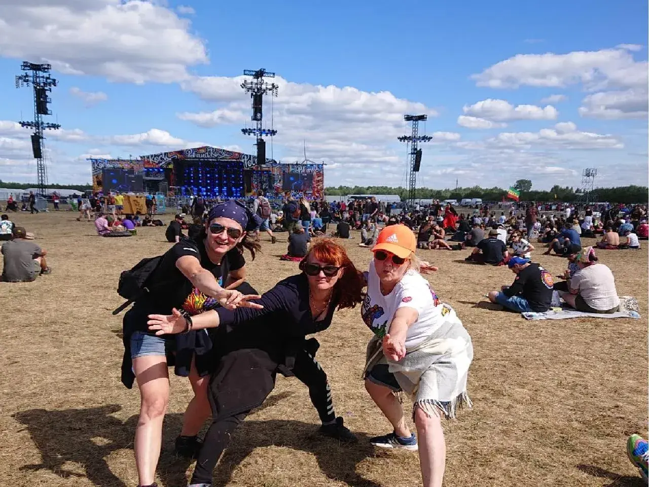 Trzy kobiety pozują na tle sceny festiwalowej Pol'and'Rock. Tłum ludzi siedzi na trawie, ciesząc się pogodą i oczekując na występ.
