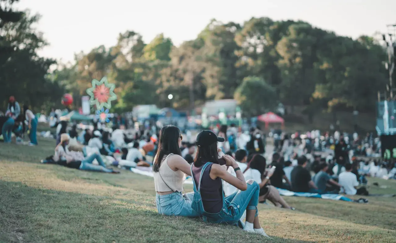 Dwie dziewczyny siedzą na trawie, ciesząc się atmosferą festiwalu. W tle widać tłum ludzi i kolorowe dekoracje, zapowiadające wspaniałe igo koncerty.