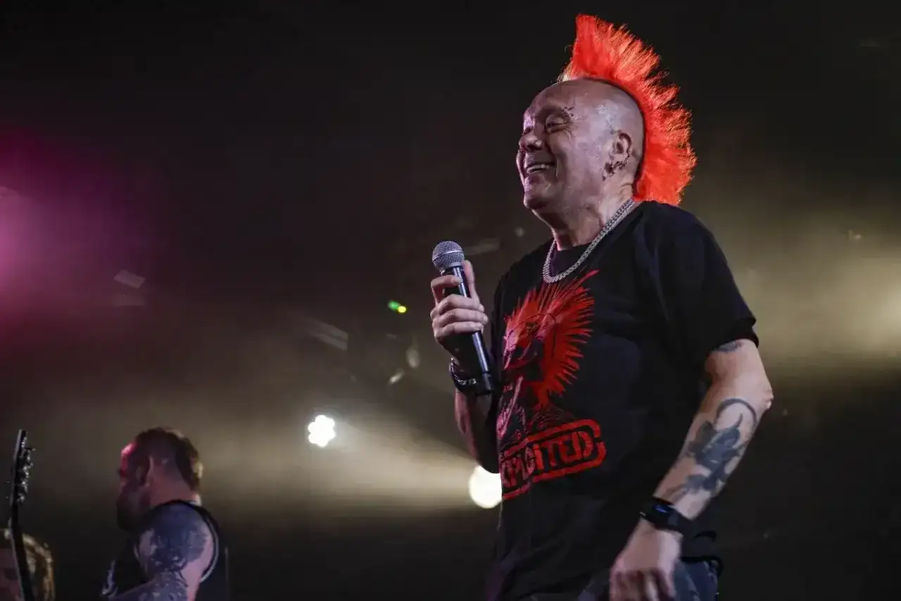 Wokalista punk rockowy z irokezem śmieje się do mikrofonu. Na koszulce ma czerwony nadruk.