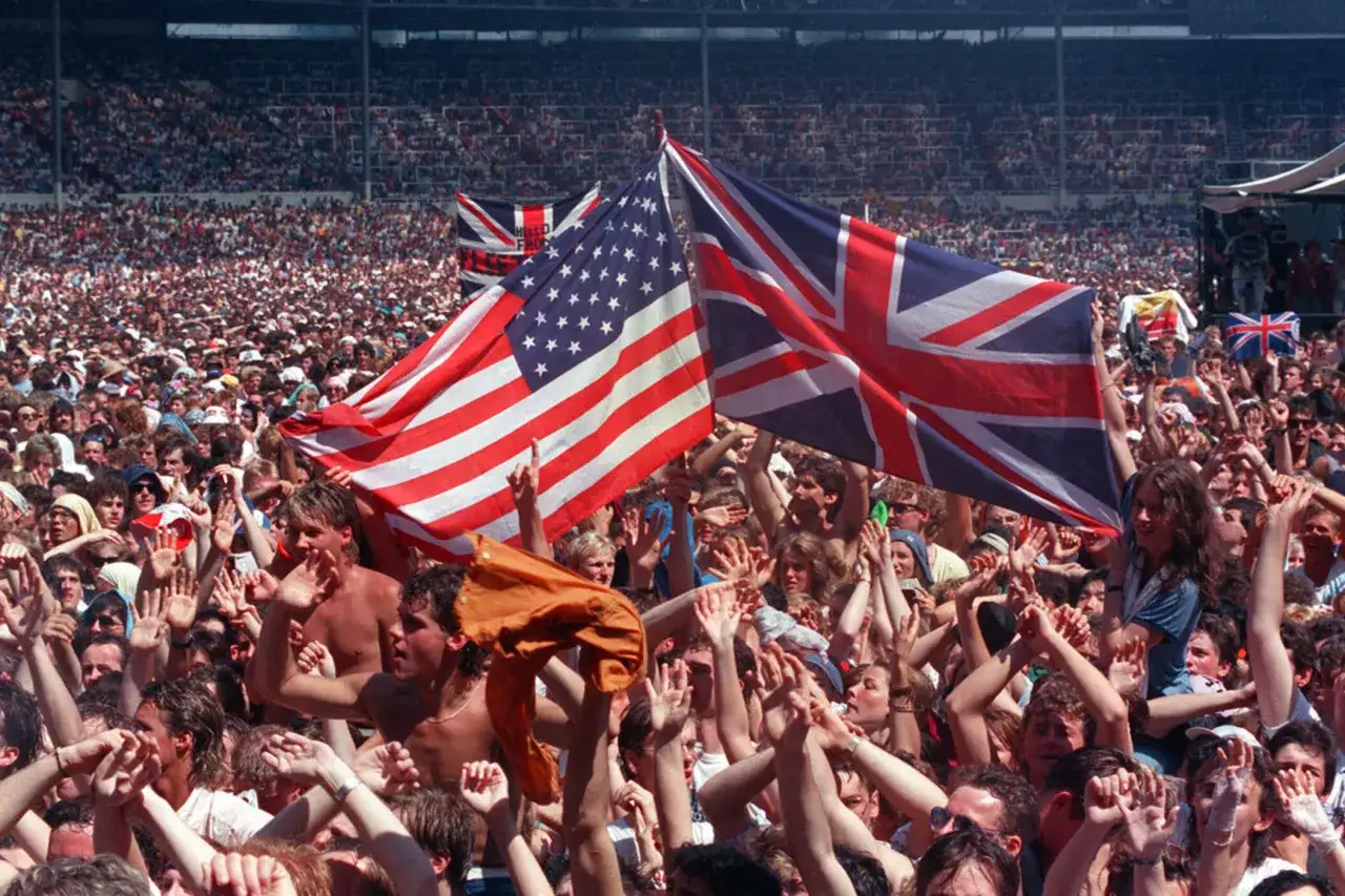 Tłum na koncercie Live Aid unosi flagi USA i Wielkiej Brytanii, celebrując muzykę i jedność.